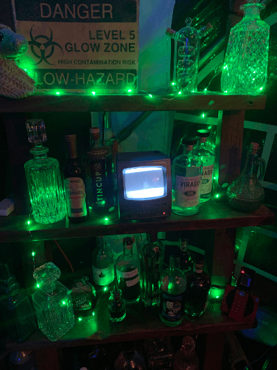 Tiny screen surrounded by bottles glowing green with sign reading Danger Level 5 Glow Zone